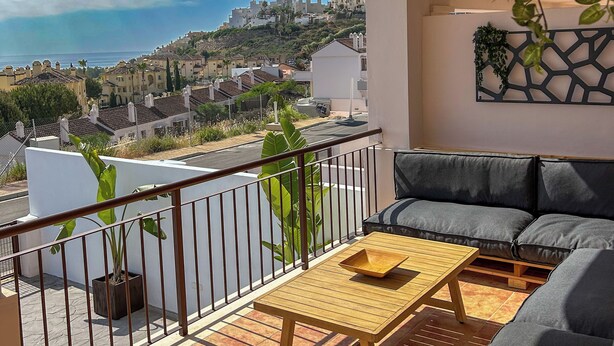 A balcony with a sofa, a wooden table, and a view of a coastal town. - Green Oasis Costa Casa Adosada con Vistas al Mar (Casares)