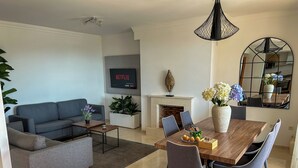 A modern living room with a grey sofa, a wooden dining table with chairs, a TV mounted on the wall, and a fireplace. - Green Oasis Costa Casa Adosada con Vistas al Mar (Casares)