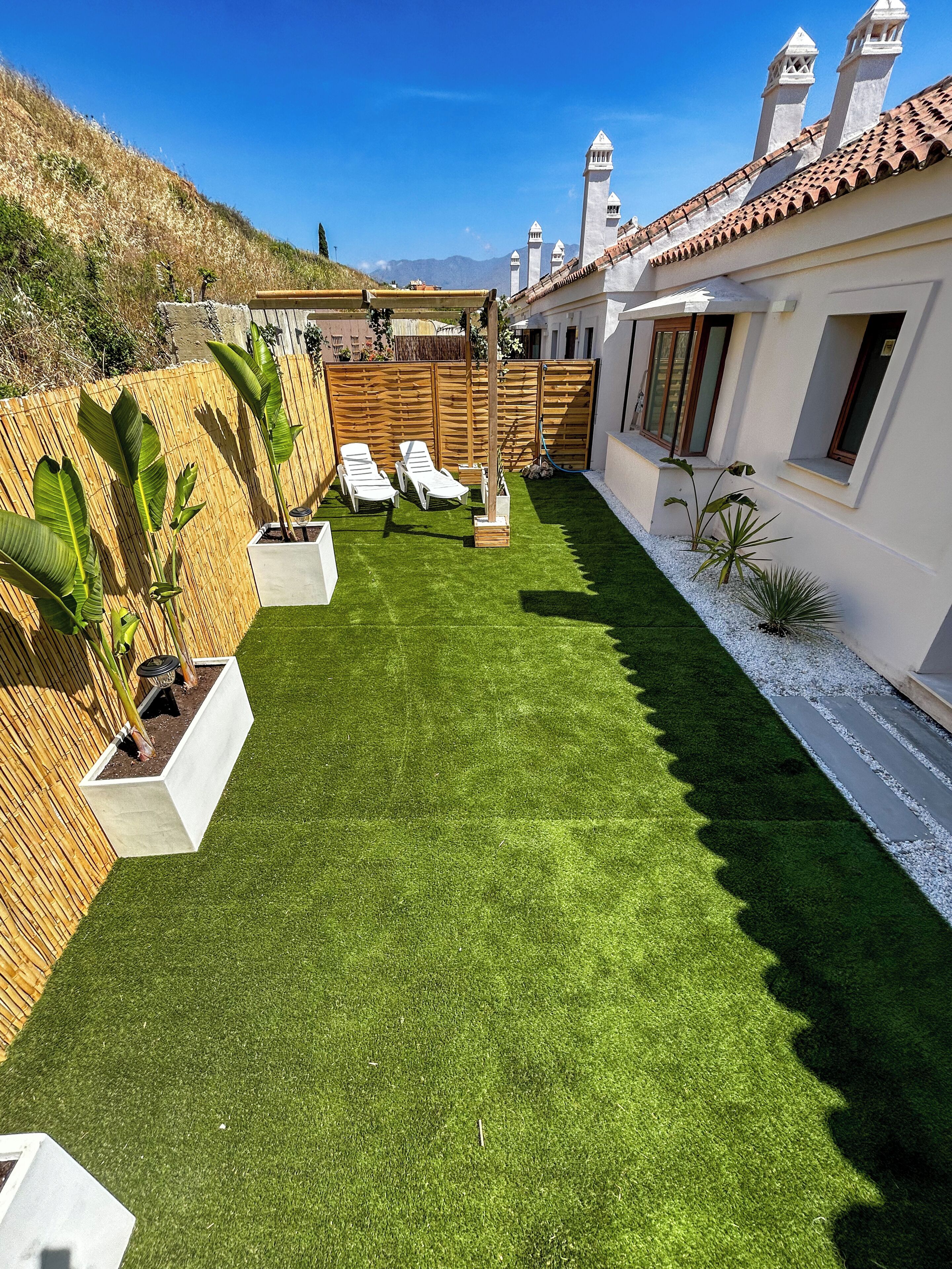 A well-maintained backyard with artificial grass, white lounge chairs, and potted plants.