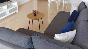 Ein modernes Wohnzimmer mit einem grauen Sofa, einem kleinen runden Tisch mit einer Blume, einer weißen TV-Konsole, einem Essbereich mit Tisch und Stühlen sowie einem Deckenventilator.