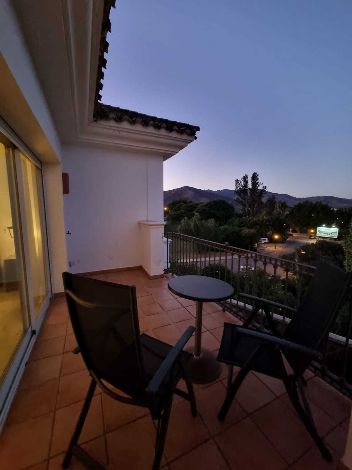 A balcony with two chairs and a small table, overlooking a road and distant mountains.
