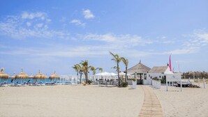 Un resort frente a la playa con sillas de descanso, sombrillas de paja y un edificio blanco con una bandera rosa.