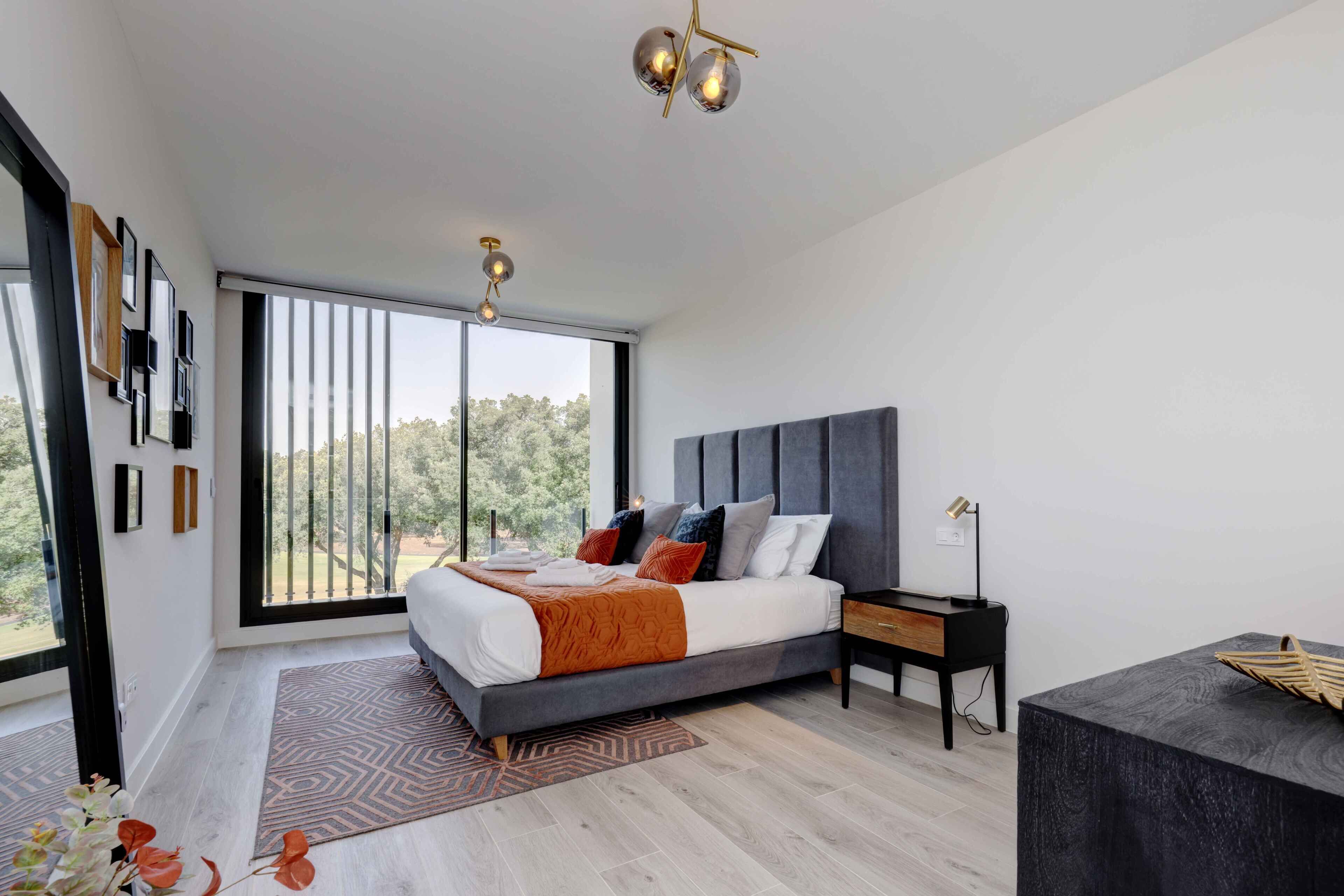 Une chambre moderne avec un grand lit, une table de chevet, un bureau et une vue sur les arbres par la fenêtre.