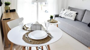 Un salon moderne avec un coin repas. Il comprend un canapé gris orné d'un coussin sur lequel est écrit « bonjour », une petite table avec une assiette et un verre blancs, et une table d’appoint en bois avec une horloge et une plante.