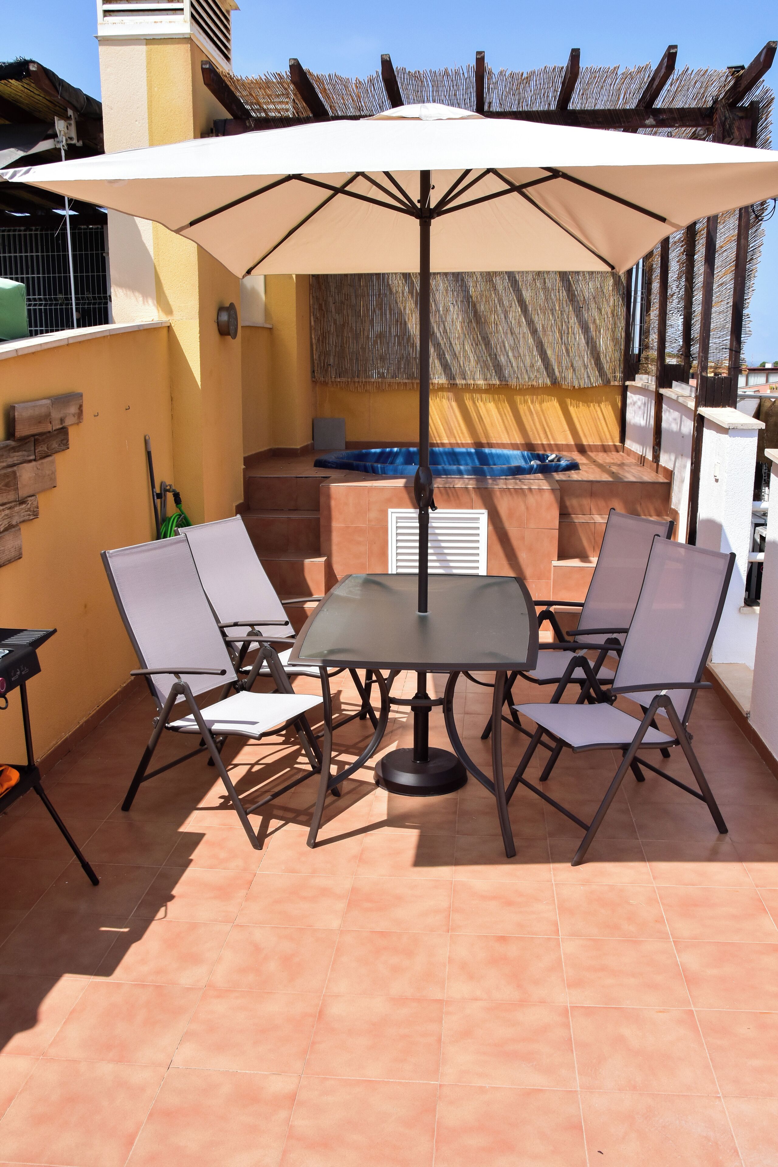 Un patio al aire libre con una mesa y cuatro sillas, un paraguas grande y un jacuzzi al fondo.
