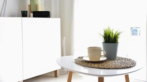 Une table blanche avec un set de table en fibres tressées, un pot gris contenant une plante verte, et une tasse et une soucoupe de couleur beige.