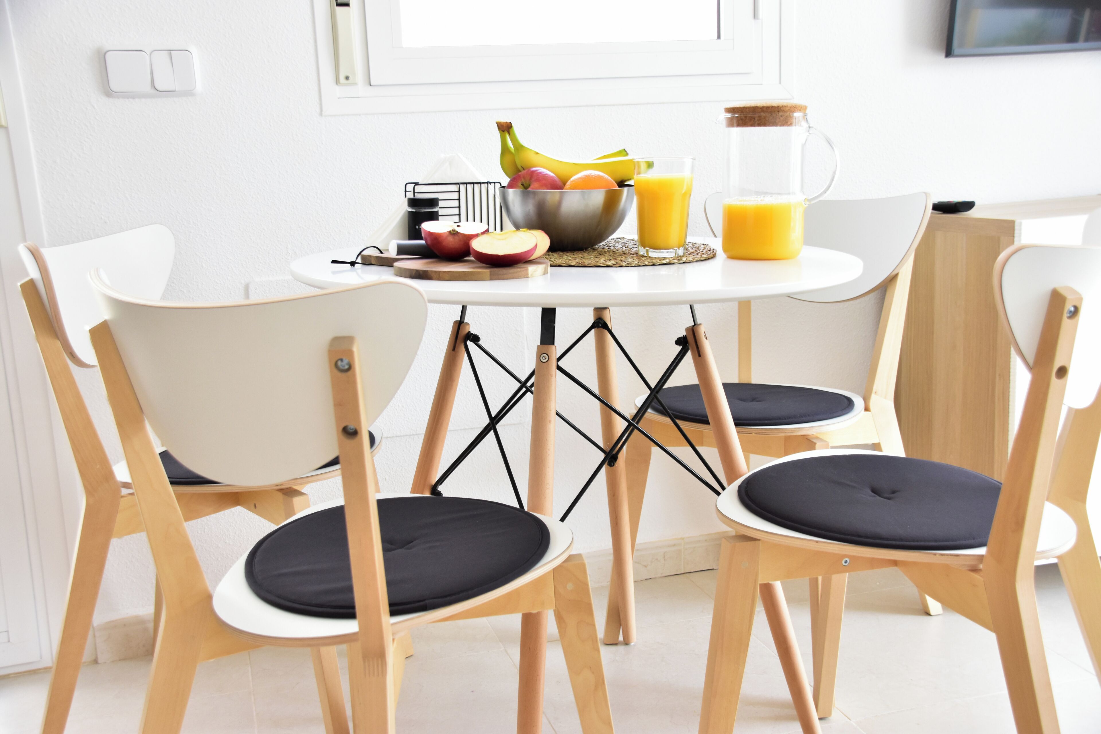 A breakfast table with a bowl of fruit, a glass of orange juice, and a pitcher. There are four wooden chairs with black cushions around the table.