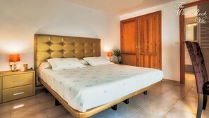Une chambre à coucher avec une tête de lit capitonnée, un lit avec une literie à motifs floraux, une table de chevet avec une lampe et une armoire en bois.
