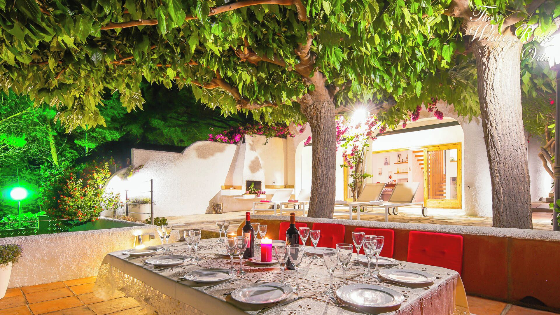 Un área de comedor al aire libre con una mesa lista para una comida, rodeada de árboles y una pared blanca.