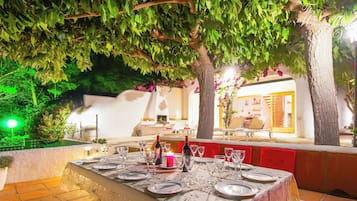 Un área de comedor al aire libre con una mesa lista para una comida, rodeada de árboles y una pared blanca.