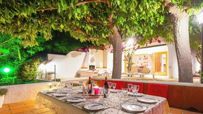 Un área de comedor al aire libre con una mesa lista para una comida, rodeada de árboles y una pared blanca.
