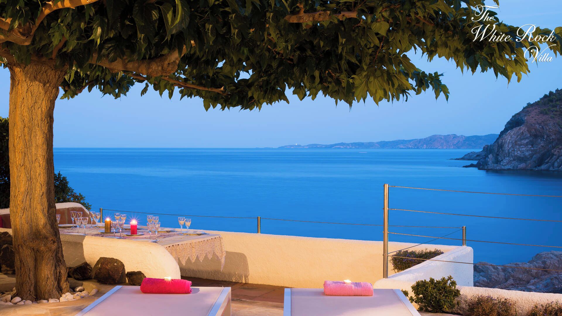 Una terraza con vistas al mar, una mesa dispuesta con vasos y velas, y un árbol en primer plano.
