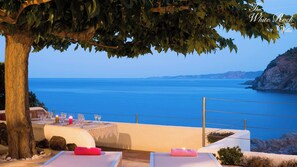 Una terraza con vistas al mar, una mesa dispuesta con vasos y velas, y un árbol en primer plano.