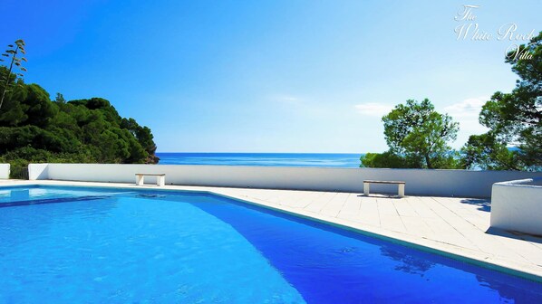 Piscina azul cristalina con vistas al mar, rodeada de muros blancos y bancos.