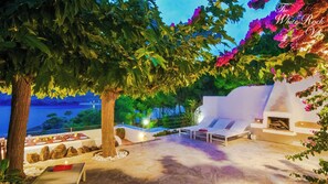 Un área de comedor privada al aire libre con vista al mar, un árbol grande y una pared blanca con chimenea.