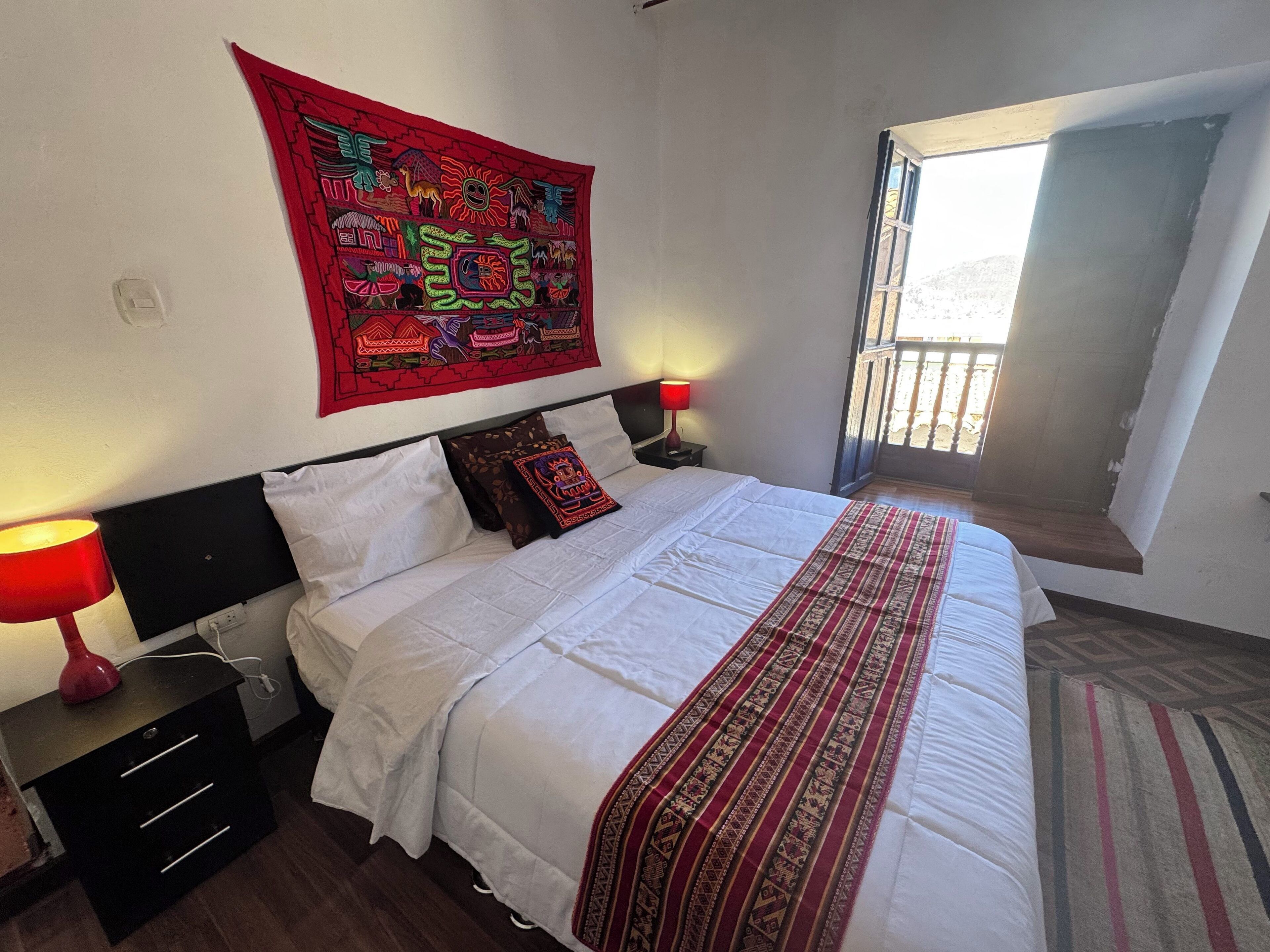 A neatly made bed with white bedding and a red patterned throw, a red lamp on a bedside table, and a colorful tapestry on the wall.