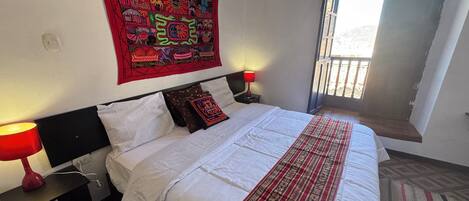 A neatly made bed with white bedding and a red patterned throw, a red lamp on a bedside table, and a colorful tapestry on the wall.