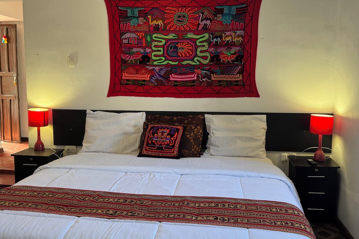 A neatly made bed with white linens and a patterned throw, flanked by two bedside tables with lamps, and a vibrant tapestry on the wall.