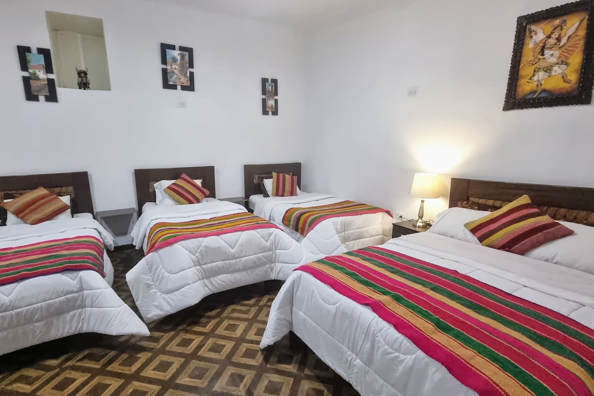 A hotel room with four beds, each with a striped blanket. There are wall-mounted paintings and a lamp on the right side.