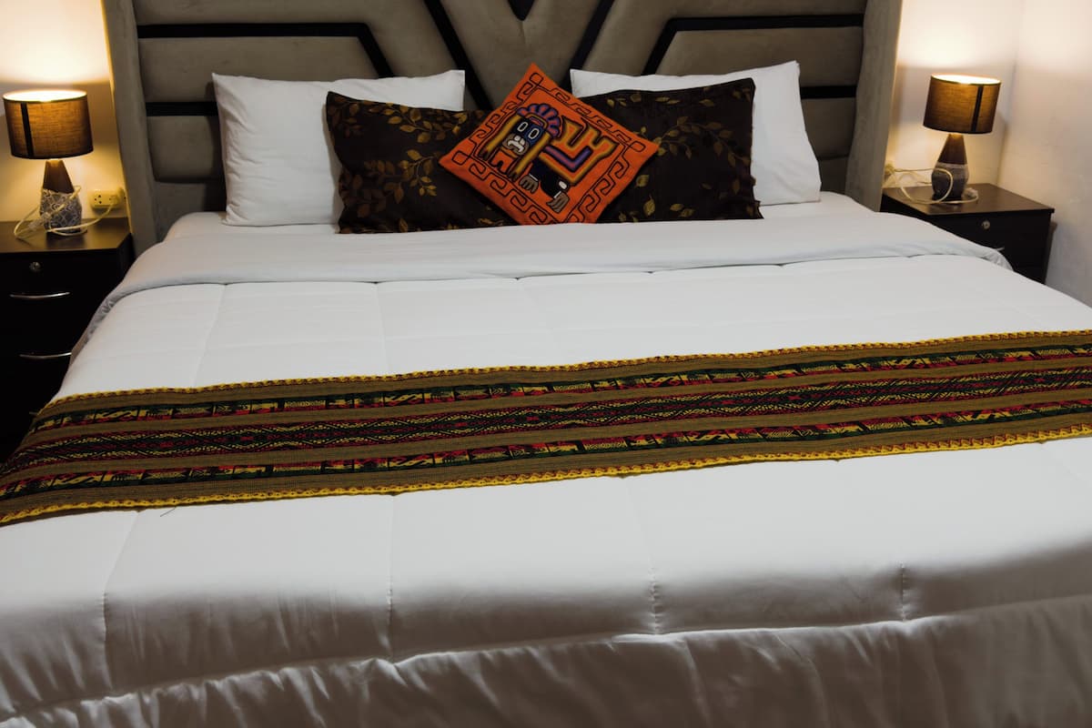 A neatly made bed with white linens, a decorative throw blanket, and a unique pillow with a geometric pattern.
