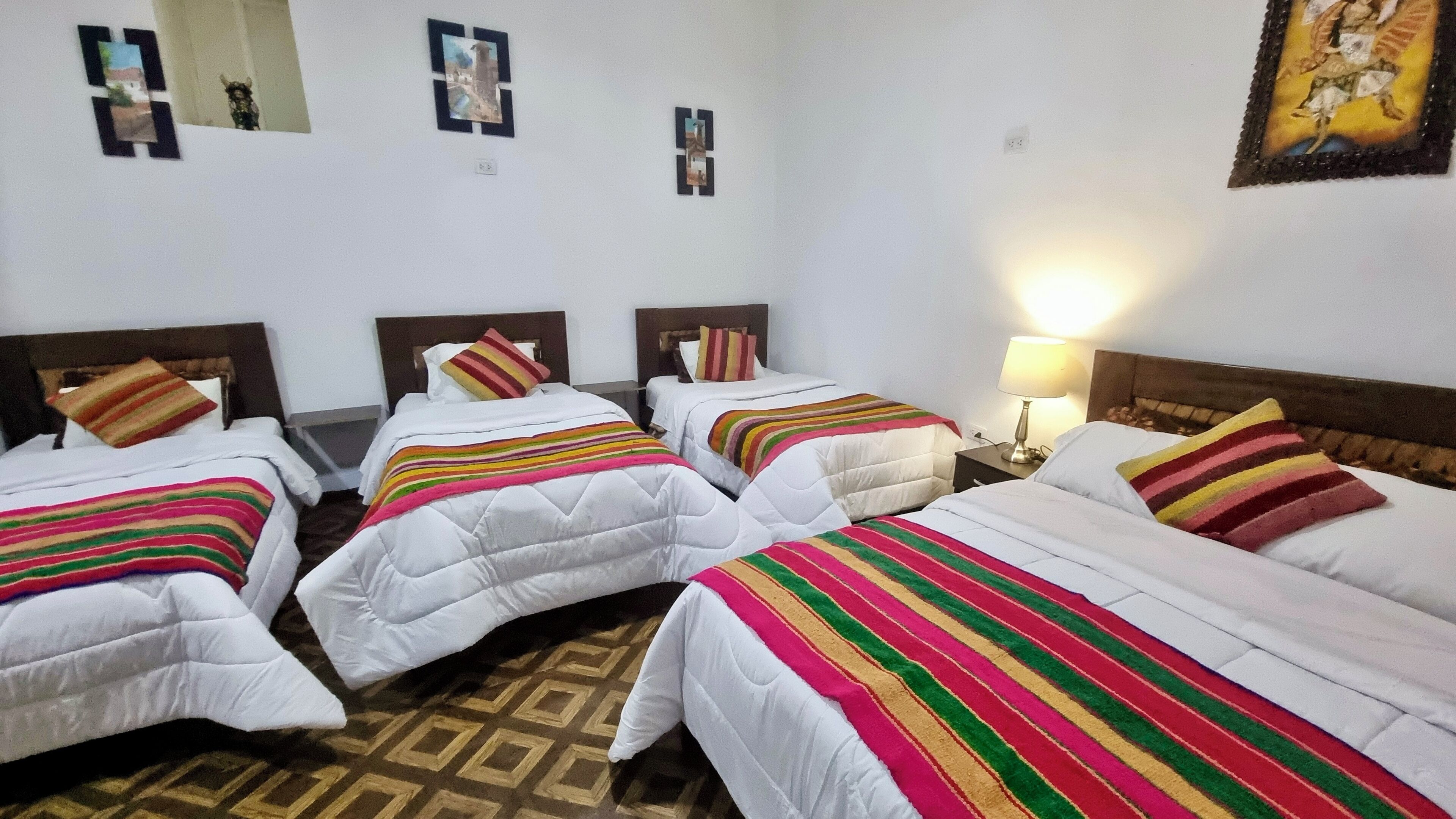 A hotel room with four single beds, each with a striped blanket and a pillow. The room has a patterned floor and a wall with framed pictures.