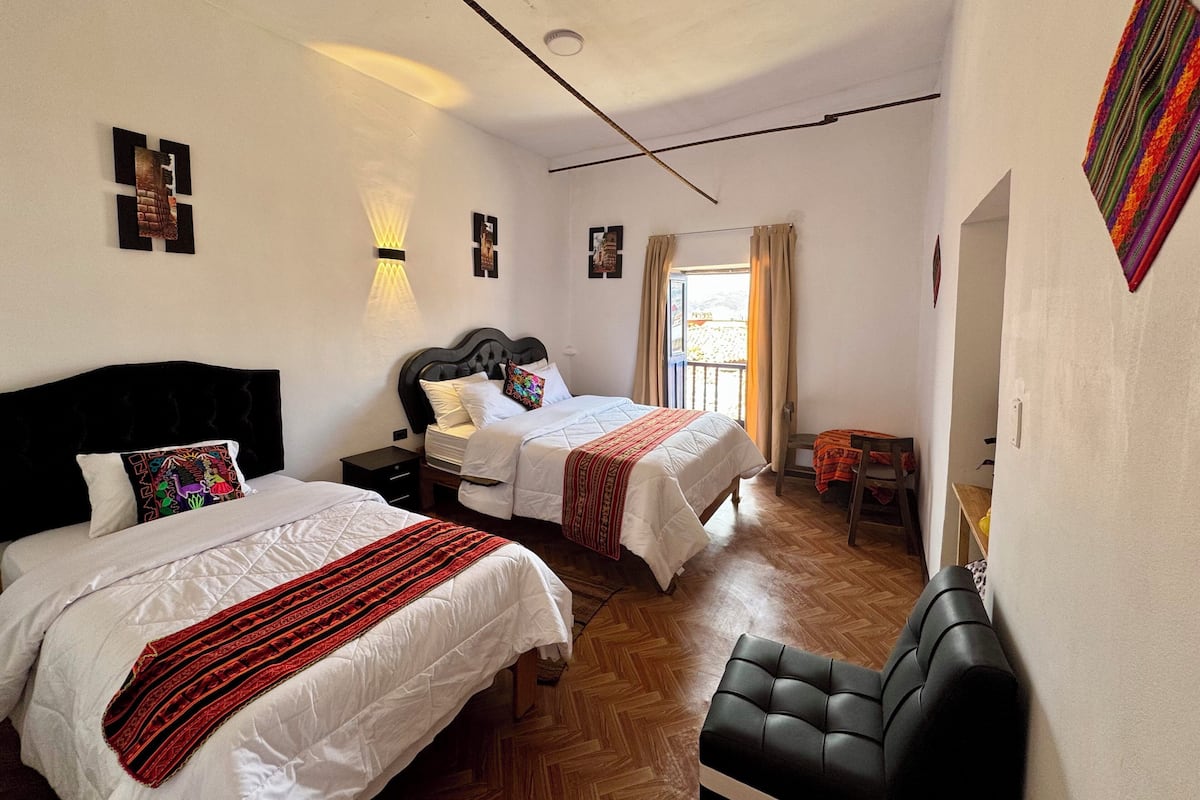 A hotel room with two beds, a black leather sofa, wooden flooring, and a balcony with curtains.