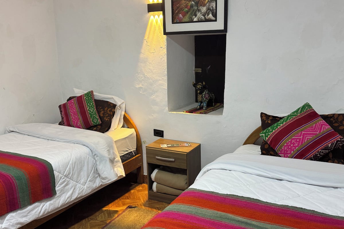 A small, cozy bedroom with two single beds, a nightstand, a wall-mounted shelf with a decorative item, and a colorful striped rug.