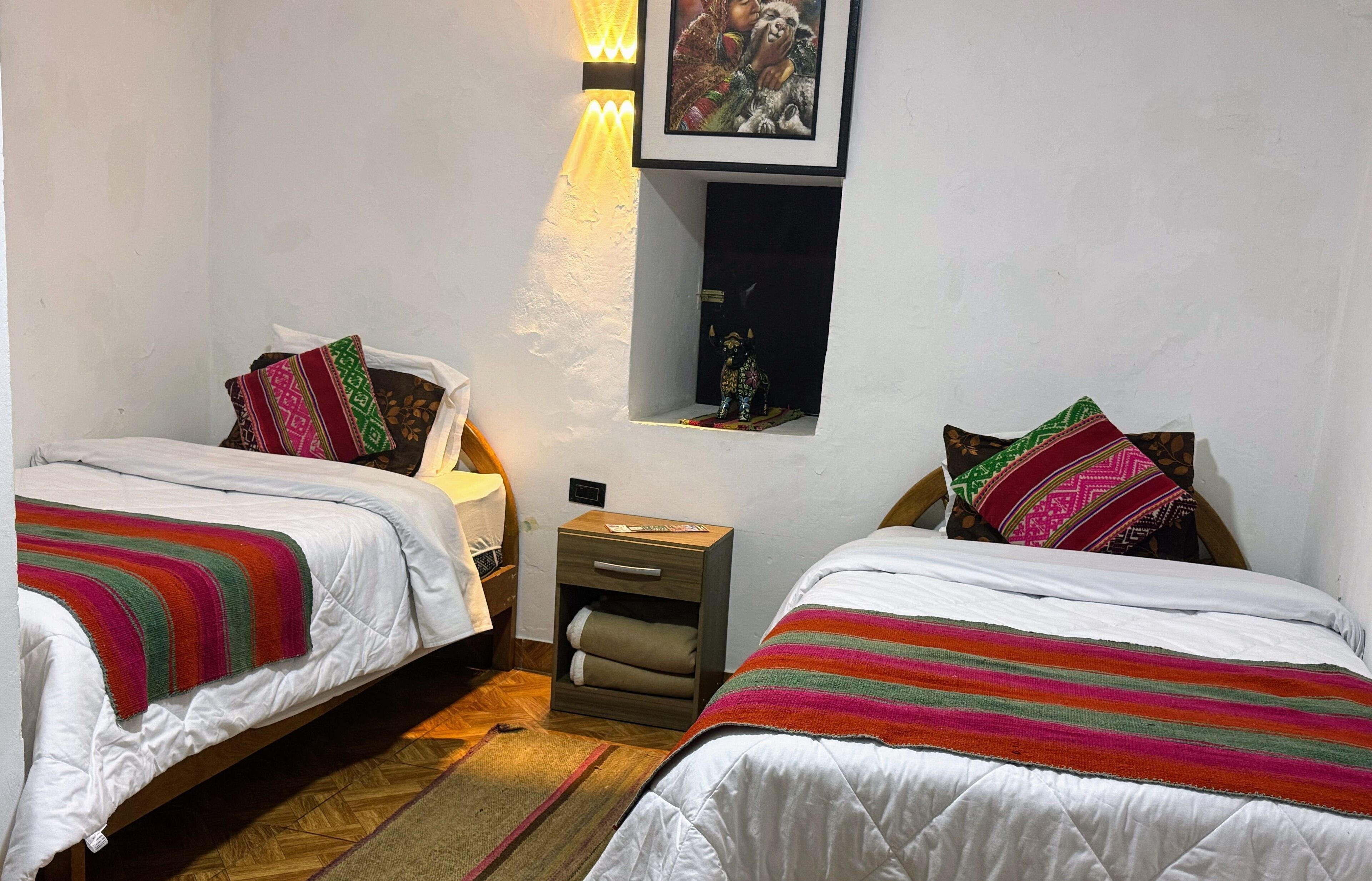 A room with two beds, each with a colorful striped blanket, a nightstand with a lamp, and a small shelf with a decorative item.
