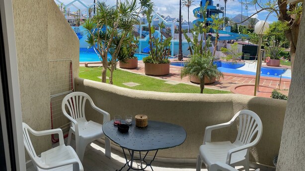 Un balcone con tavolo e sedie, vista su un'area piscina con palme e uno scivolo acquatico blu.