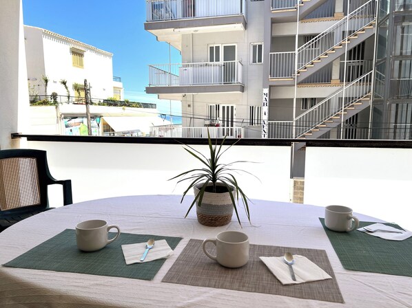 Un balcón con una mesa preparada para dos personas, una planta en maceta y vista a un edificio moderno.