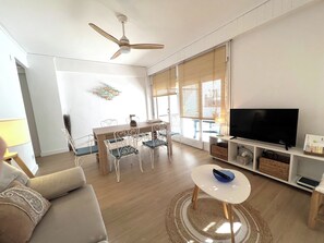 Una sala de estar con un sofá, un televisor, una mesa de comedor con sillas, un ventilador de techo y ventanas amplias.