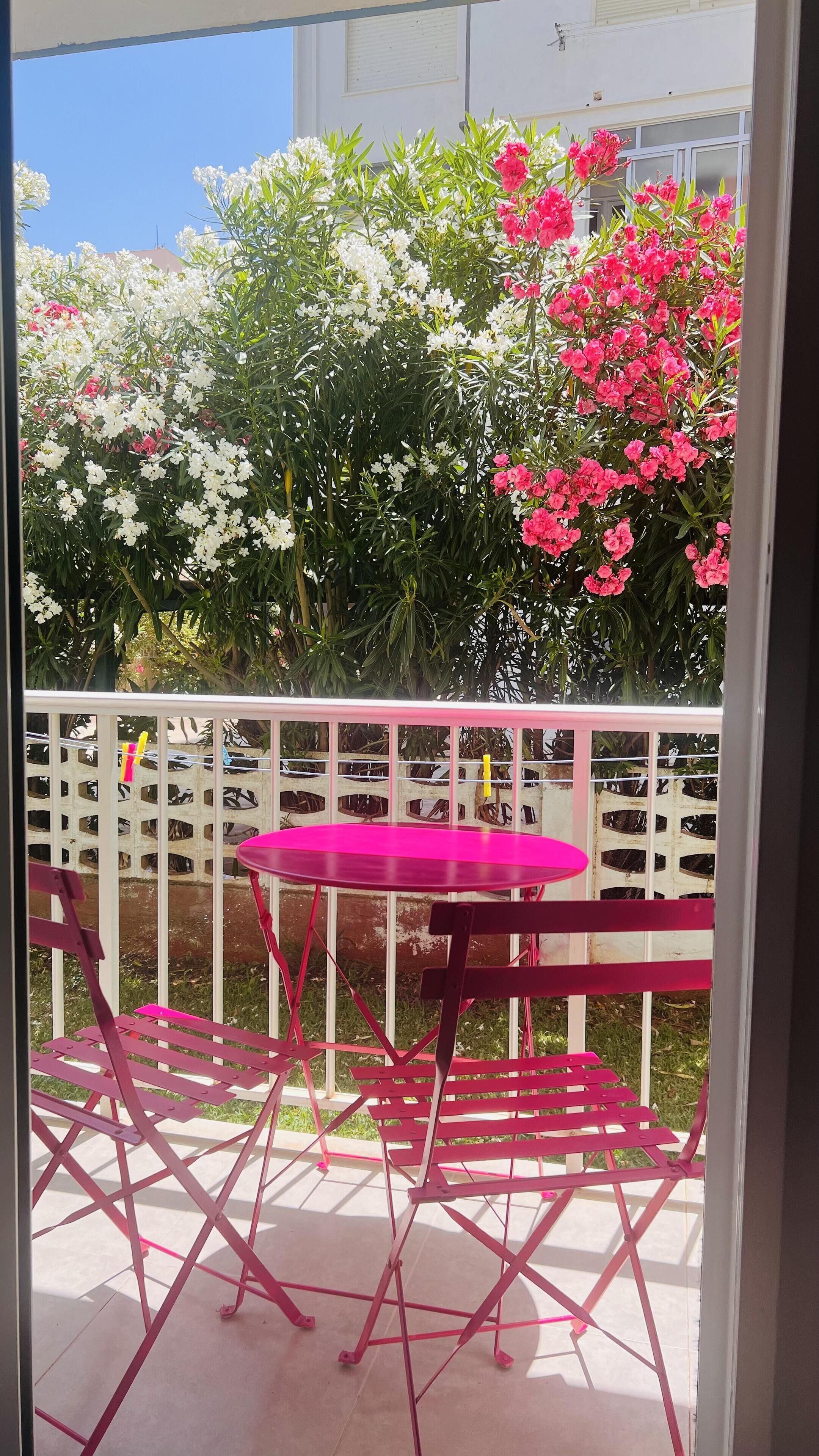 A balcony with a pink metal table and chairs, surrounded by a white fence and various flowering plants.