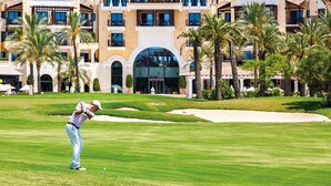 A golf course with a player in mid-swing, palm trees, and a hotel with multiple balconies in the background. - Villa Fresno With Pool on Mar Menor Golf Resort (Torre-Pacheco)