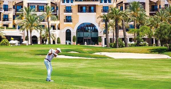 Un campo de golf con un jugador en pleno balanceo, palmeras y un hotel con varios balcones al fondo.