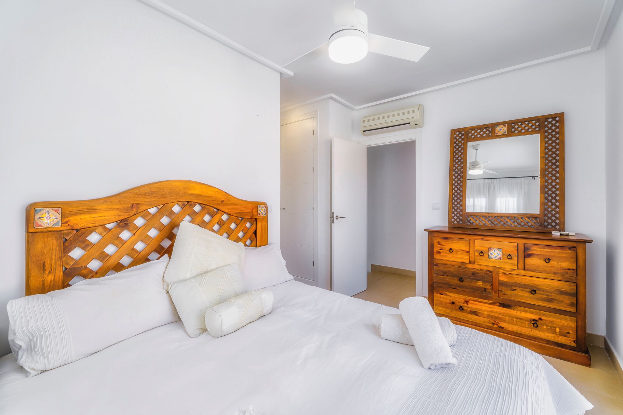 Een slaapkamer met een houten hoofdbord, een wit bed, een houten dressoir en een plafondventilator.