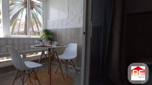 Een balkon met een tafel en twee stoelen, uitzicht op palmbomen, en een raam met een plant op de vensterbank.