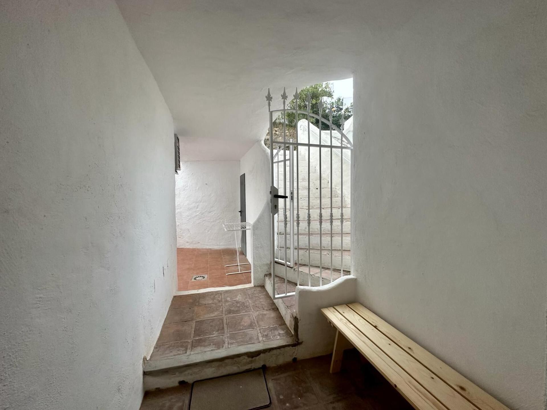 A narrow corridor with a bench, a metal gate, and a door leading to an outdoor area with a tiled floor.