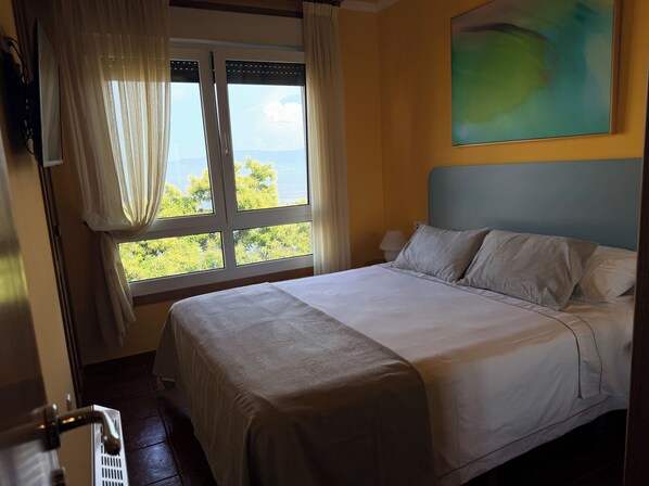 Une chambre d’hôtel avec un lit, une fenêtre avec des rideaux, un tableau au mur et une vue sur la verdure et l’eau à l’extérieur.