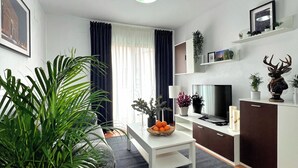 Ein modernes Wohnzimmer mit Sofa, Fernseher und Couchtisch. Pflanzen, ein Bücherregal und ein Schrank sind ebenfalls vorhanden.