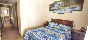 A bedroom with a bed, a nightstand, a lamp, and a painting of a waterfall on the wall. - Comfortable 2-bed Apartment in Costa Esuri (Ayamonte)