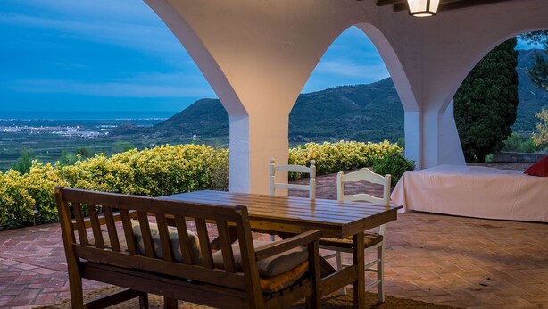 A covered outdoor seating area with a table and benches, overlooking a scenic landscape with hills and a distant town. - House Sleeps 8 - Natural Pool, Pet Friendly, Views (Nules)