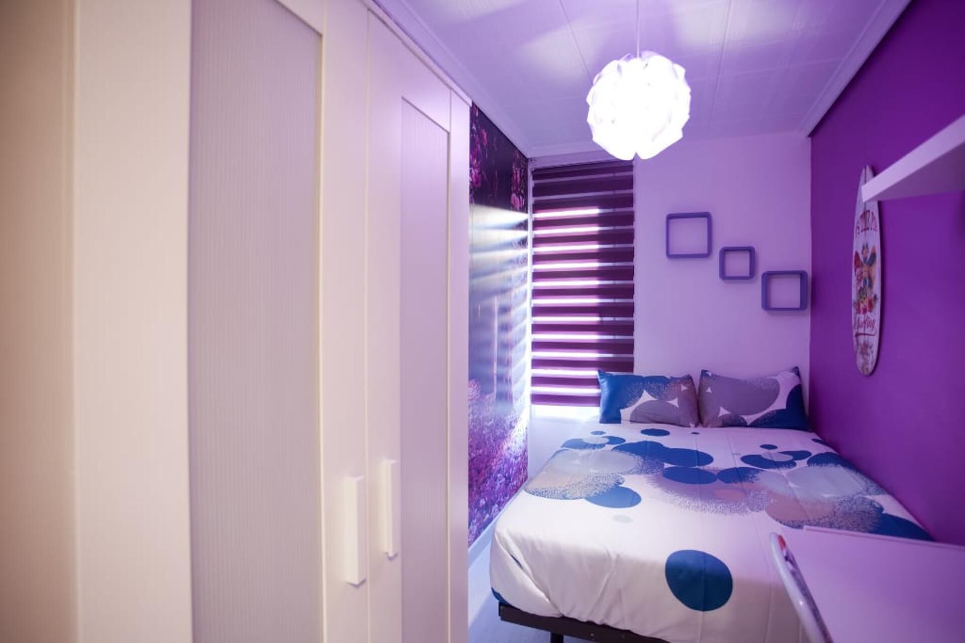 A bedroom with a bed, a window with blinds, a wall with framed pictures, and a ceiling light fixture.