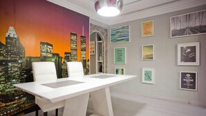 Ein moderner Arbeitsbereich mit einem weißen Konferenztisch, zwei weißen Stühlen, einem Stadtbild-Wandgemälde und verschiedenen gerahmten Kunstwerken an den Wänden.