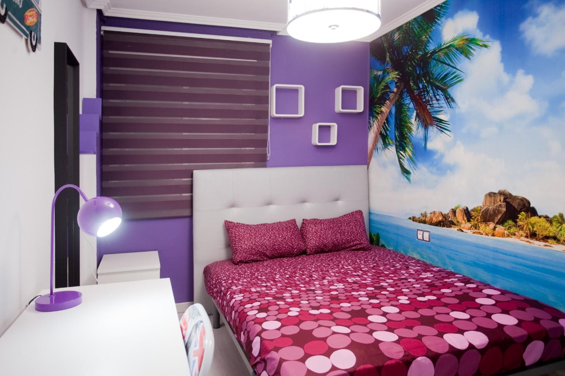Une chambre à coucher comprenant un lit, une table de chevet, une lampe, une fenêtre avec des stores et une fresque murale représentant une scène de plage.