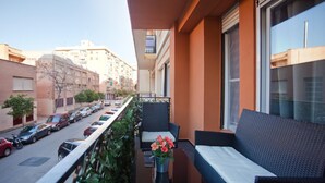 Ein Balkon mit Korbmöbeln, einem kleinen Tisch mit Blumenvase und Blick auf eine Straße mit geparkten Autos und Gebäuden.