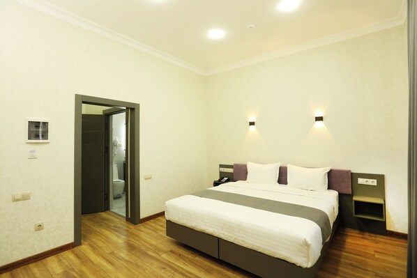 A hotel room with a large bed, a bedside table, a nightstand, a wall-mounted light, and a door leading to a bathroom. - Parklane Hotel (Tashkent)