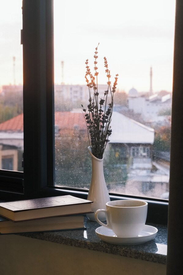 Una ventana con una taza y platillo blancos, un florero con flores secas y un libro en el alféizar.