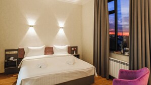 A hotel room with a large bed, two bedside lamps, a nightstand with a phone, a window with curtains, and a purple chair. - Parklane Hotel (Tashkent)