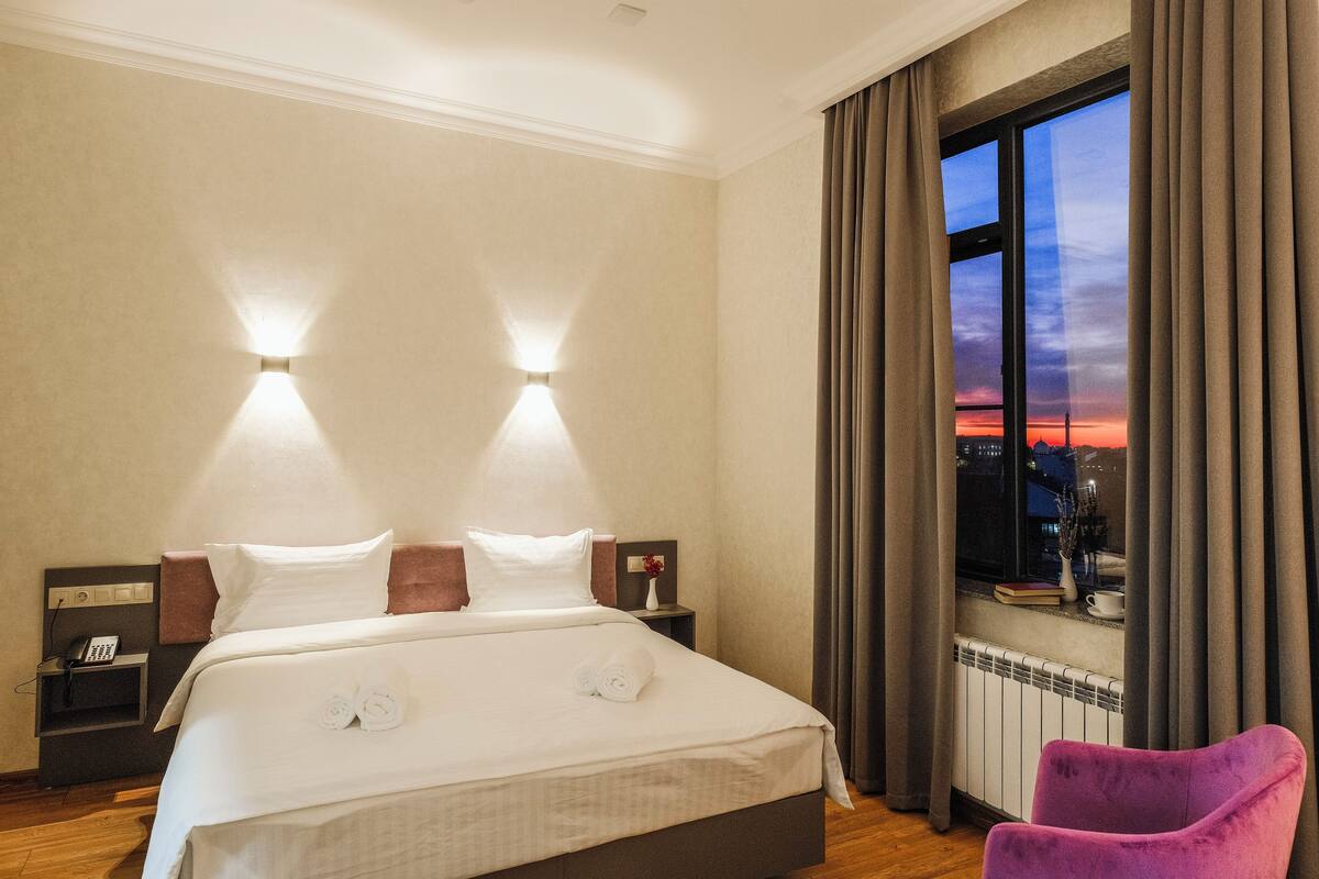 A hotel room with a large bed, two bedside lamps, a nightstand with a phone, a window with curtains, and a purple chair.
