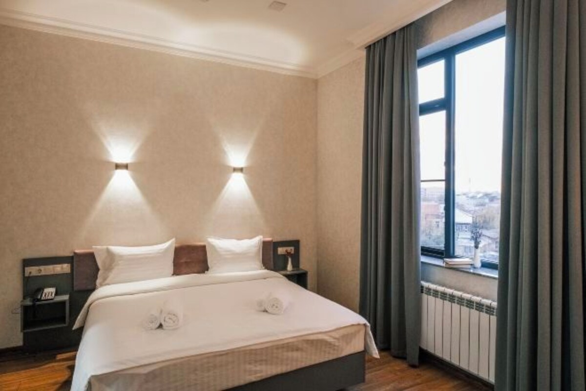A hotel room with a bed, two bedside lamps, a window with curtains, and a view of the city.